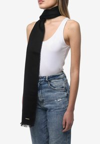 Écharpe noire en tissu doux, aux bords frangés, portée autour du cou. Modèle portant un débardeur blanc et un jean bleu clair.