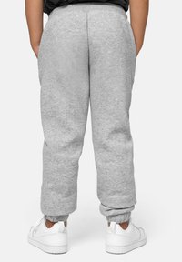 Persona vestida con pantalones de chándal grises y zapatillas blancas, de pie con las manos en los bolsillos, vista desde atrás contra un fondo liso.
