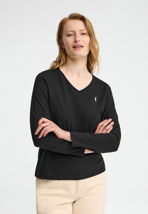 Kobieta z jasnoczerwonymi włosami w czarnej, długorękawowej koszulce z dekoltem w serek z logo i beżowych spodniach, z założonymi rękami, na białym tle.