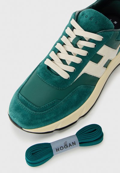 Green and white suede sneaker with beige laces next to a pair of matching green replacement shoelaces branded Hogan on a white background.