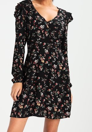 Femme portant une robe noire à manches longues imprimée fleurie avec un col en V et des détails à volants aux épaules.