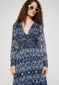 Rochie lungă, cu un model albastru, cu un decolteu adânc în V și mâneci lungi din material transparent. Dispune de o talie elastică și accente texturate. O pereche de ochelari de soare albi.