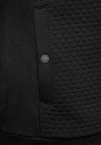 Černá mikina s texturovaným vzorem na jedné straně, s hladkým materiálem na druhé straně a malým logem na kapse.