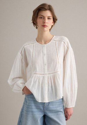 Mujer con cabello corto lleva una blusa blanca con botones, detalles de encaje y mangas largas abullonadas, combinada con jeans azul claro.