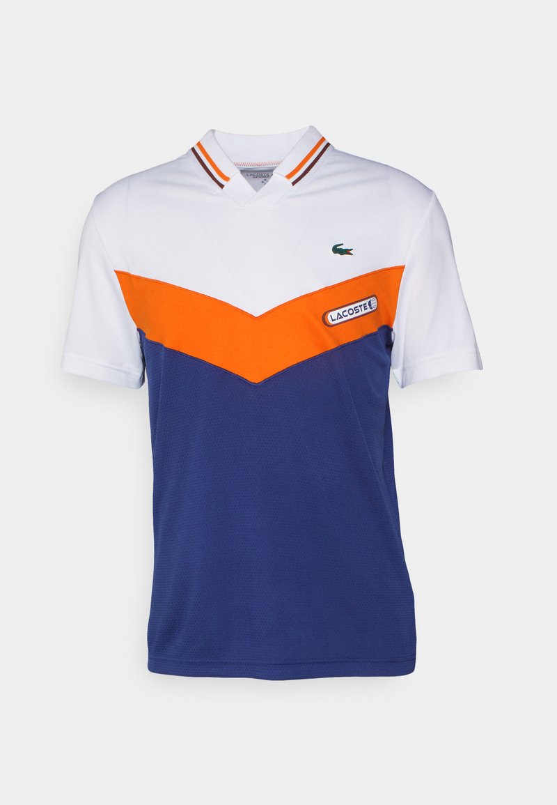 Polo blanc et marine avec un design en chevron orange, un col en ribbed avec des rayures orange et marine, et un logo Lacoste sur la poitrine.