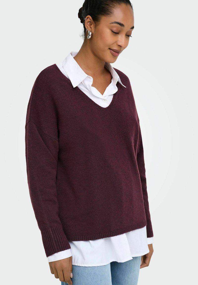 Femme aux cheveux foncés en chignon, portant des boucles d’oreilles créoles argentées, une chemise blanche à col sous un pull ample couleur bordeaux, et un jean bleu clair.