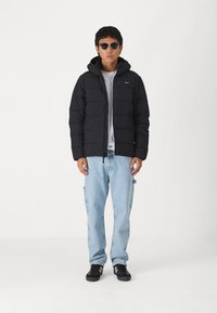 Svart puffjacka med huva som har quiltade sektioner, bärs över en grå t-shirt, ihop med ljusblå jeans och svarta sneakers.