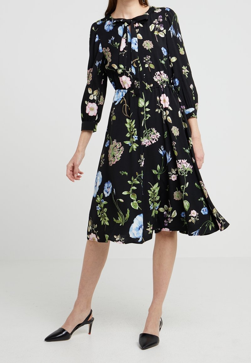 Femme portant une robe midi noire à imprimé floral multicolore, manches trois-quarts, col noué, et escarpins noirs à bride arrière pointus.