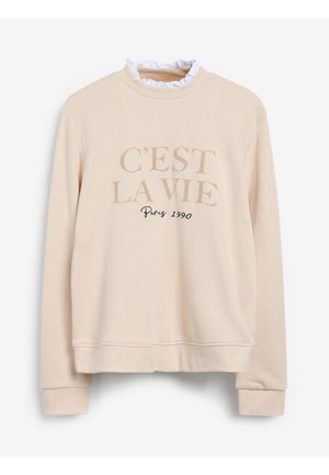 Femme portant un sweat-shirt beige avec texte et un pantalon en cuir noir cintré, mains dans les poches, baskets blanches.