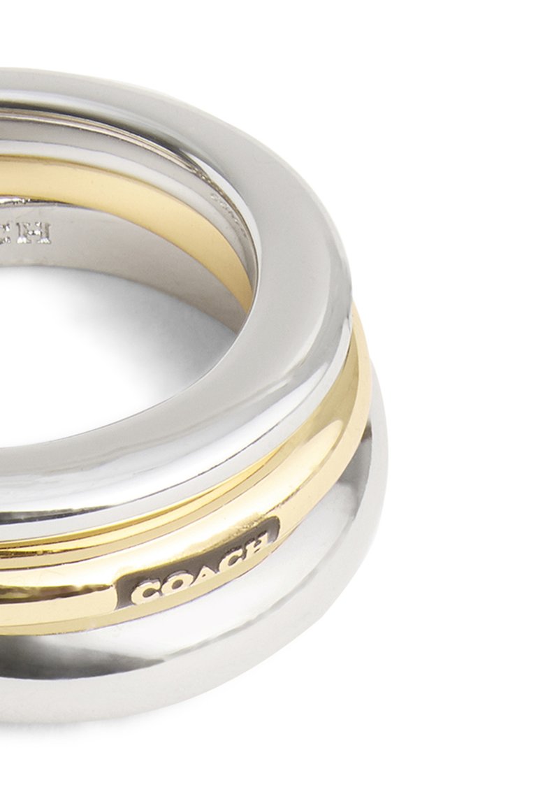 Anillo en tonos plateado y dorado con un diseño redondeado, que presenta acabados suaves y un logo de "COACH" en la banda. Varias capas de metal se entrelazan.