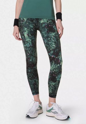 Vrouw die groene en zwarte legging met patroon draagt, witte en groene sportschoenen, zwarte polsbandjes en een groene top, staand met ontspannen handen.