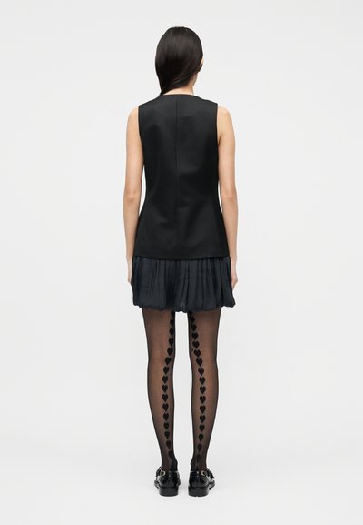 Γυναίκα με μακριά σκούρα μαλλιά φορά αμάνικο μαύρο τοπ, πλισέ μίνι φούστα, μαύρο καλσόν με κάθετο μοτίβο καρδιάς και μαύρα παπούτσια, στέκεται με την πλάτη.