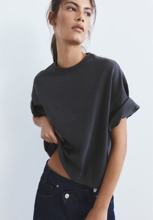 Ung kvinde løfter let en mørk oversize T-shirt og bærer mørkeblå jeans mod en ensfarvet lys baggrund.