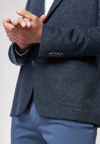 Ciemnoniebieska blazer z teksturą, posiadająca dwie przednie kieszenie i cztery guziki na mankiecie, w zestawie z jasnoniebieskimi spodniami.