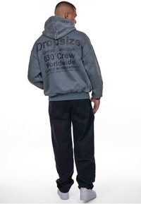 Grauer oversized Hoodie mit einer großen Kapuze und einem großen schwarzen Grafikdruck auf dem Rücken. Kombiniert mit schwarzen Hosen und weißen Sneakern.