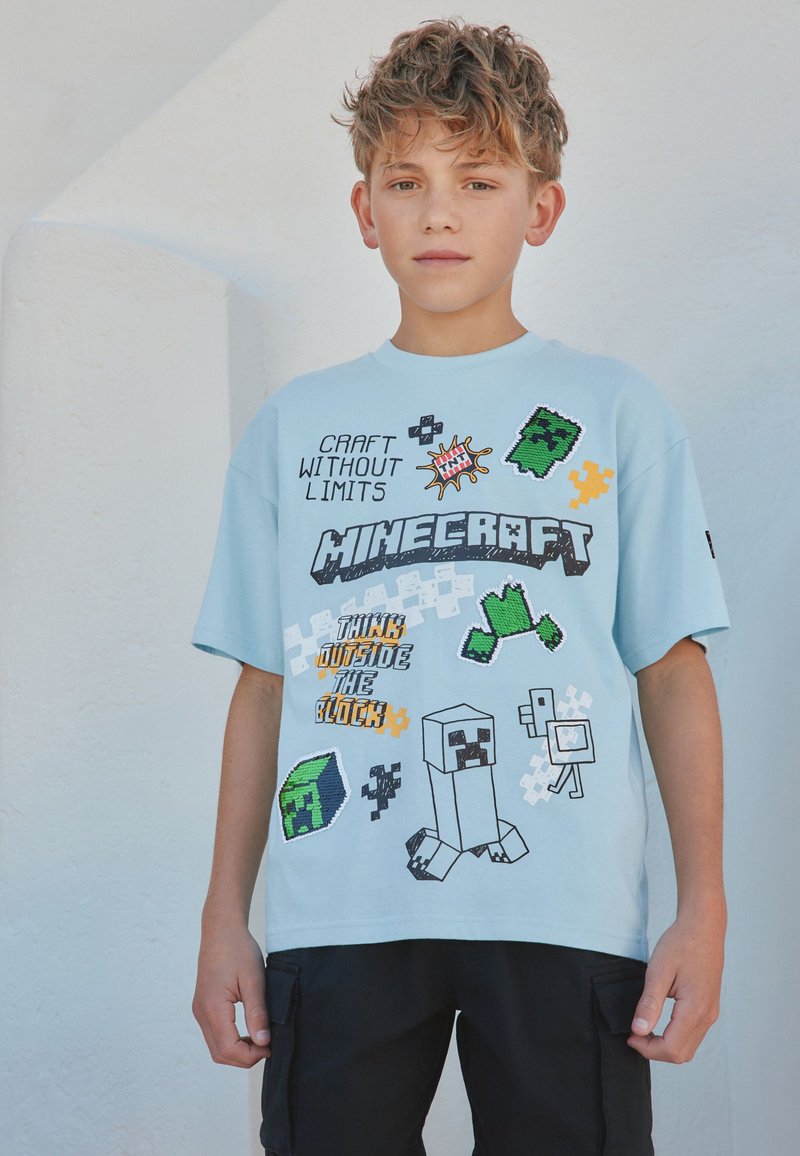 Băiat cu păr creț care poartă un tricou albastru deschis cu tematică Minecraft cu personaje pixelate și text, stând lângă un perete alb.