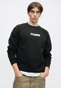 Μαύρη μπλούζα με στρογγυλή λαιμόκοψη και ριμπ ύφανση στα μανίκια, με λευκό γραφικό κείμενο στο κέντρο. Φορεμένη πάνω από ένα λευκό πουκάμισο και συνδυασμένη με μπεζ παντελόνι.