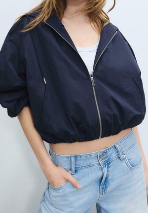 Femme portant une veste courte bleu marine zippée sur un haut blanc avec un jean taille haute bleu clair, main dans la poche, sur un fond uni.