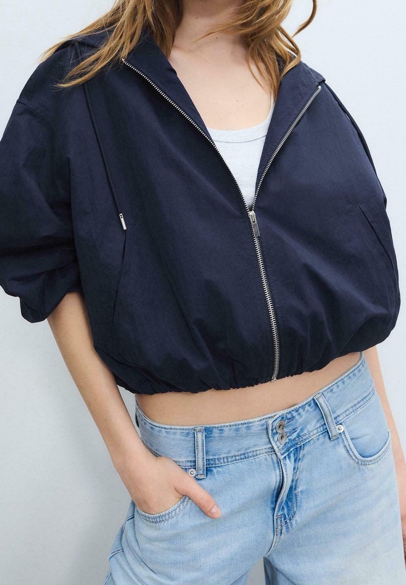 Femme portant une veste courte bleu marine zippée sur un haut blanc avec un jean taille haute bleu clair, main dans la poche, sur un fond uni.