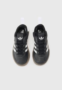 Mustat urheilukengät, joissa on kolme valkoista viivaa ja musta kieli, jossa on logo. Pohja on kuvioitu ja hiekanruskea.