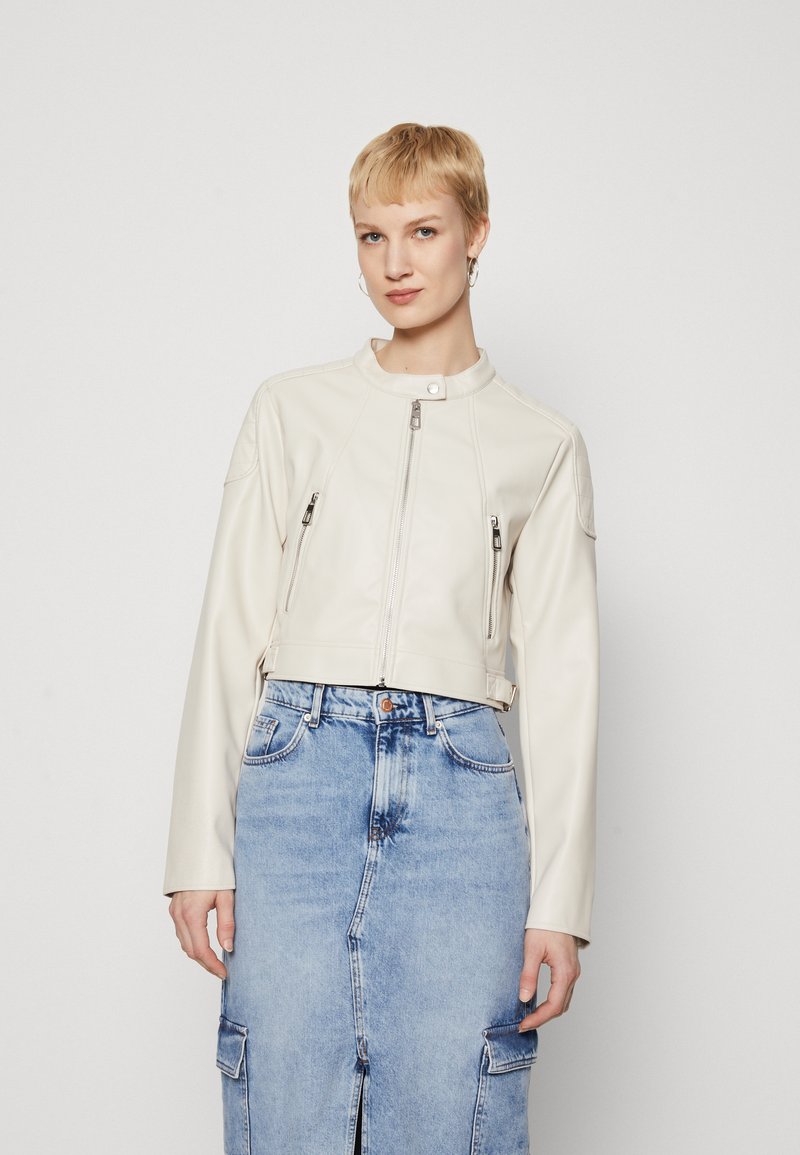 Veste cropped en cuir blanc avec un col montant, fermeture éclair à l'avant et deux poches zippées. Portée sur une jupe en jean.