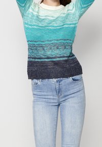 Femme portant un pull à manches longues avec un dégradé de blanc, sarcelle et bleu marine, associée à un jean en denim bleu clair, les bras levés au-dessus de la tête.