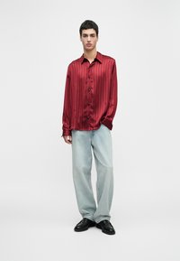 Young man standing wearing a loose red satin button-up shirt, light blue wide-leg jeans, and black loafers on a plain white background.