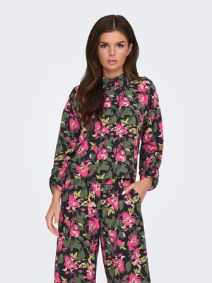 Kvinde iført matchende bluse og bukser med blomsterprint i grøn, pink og sort, stående med den ene hånd i lommen mod en ensfarvet baggrund.