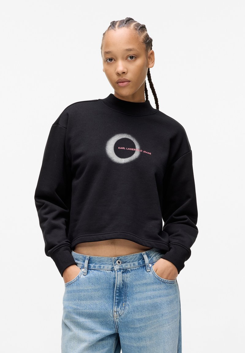 Fekete rövidített pulóver magas nyakkal, kör alakú logóval és piros "KARL LAGERFELD JEANS" felirattal. Világos mosású farmernadrággal párosítva.
