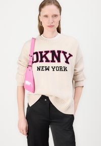 Sweater bege com letras em preto e rosa "DKNY NEW YORK", punhos canelados, combinado com calças pretas e uma bolsa rosa brilhante.