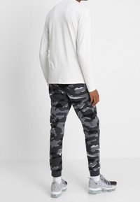 Vit långärmad topp i kombination med svart och grå kamouflagebyxor, med elastiska ärmslut och reflekterande detaljer. Silverfärgade sneakers fulländar looken.