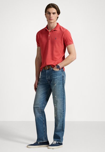 Rød polo-skjorte med korte ermer og krage, kombinert med slitte blå jeans med lapper og frynser. Modellen har på seg blå sneakers.