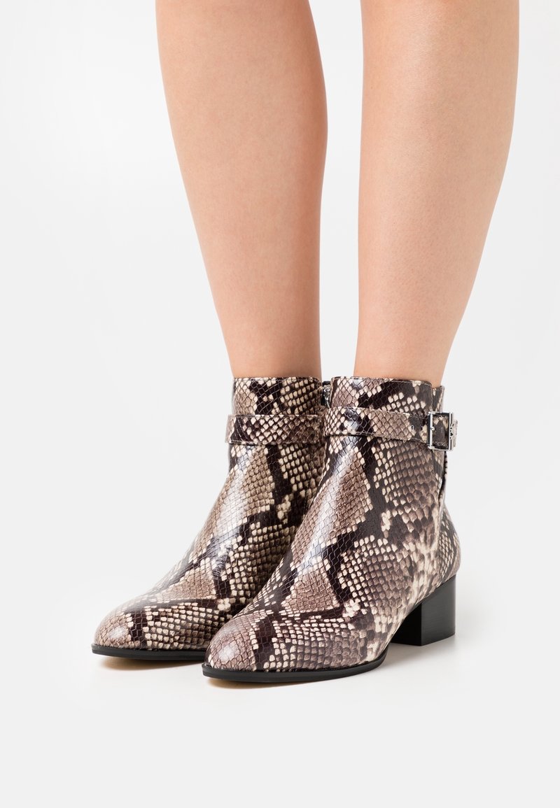 Bottes chevilles avec motif peau de serpent noir et beige, talon bas et épais, fermeture éclair latérale et sangle à boucle, portées sur jambes nues.