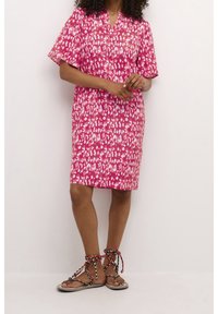 Kvinna klädd i en knälång rosafärgad mönstrad klänning och platta sandaler med röda och svarta repremmar mot en enfärgad vit bakgrund.