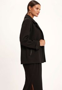 Czarna wełniana kurtka z kołnierzem, bocznymi kieszeniami na zamek i luźnym krojem, zestawiona z czarną spódnicą z prążkowanej tkaniny z bocznym rozcięciem.