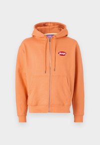 Orange zip-up hoodie made of soft fabric, featuring a front pocket, drawstring hood, and a red logo patch on the upper left side.