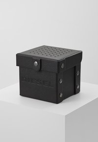 Boîte en cuir noir avec couvercle perforé, présentant des accents en métal argenté et le logo "DIESEL" en relief sur le devant. Forme carrée.