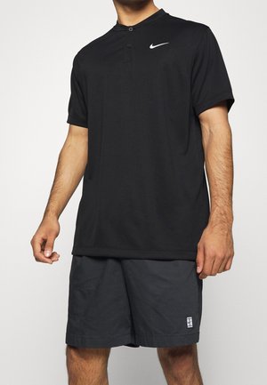 Homme portant un polo Nike noir à manches courtes et un short de sport noir, debout devant un fond uni.