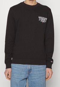 Felpa nera in cotone con design a girocollo, polsini e orlo a coste, e un logo bianco sul cuore a sinistra. Indossata con jeans blu.