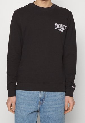 Sudadera de algodón negra con diseño de cuello redondo, puños y dobladillo acanalados, y un logo blanco en el lado izquierdo del pecho. Combinada con jeans azules.