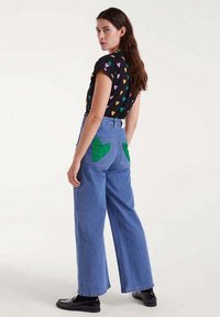 Mujer vistiendo una camiseta negra con corazones coloridos y jeans anchos azules con parches de corazones verdes en los bolsillos traseros, de pie, mirando por encima del hombro.
