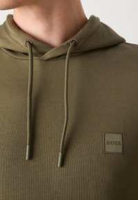 Olijfgroene hoodie met een geribbelde textuur, trekkoorden en een vierkante logo-patch op de borst. Heeft een klassiek ontwerp met een ontspannen pasvorm.