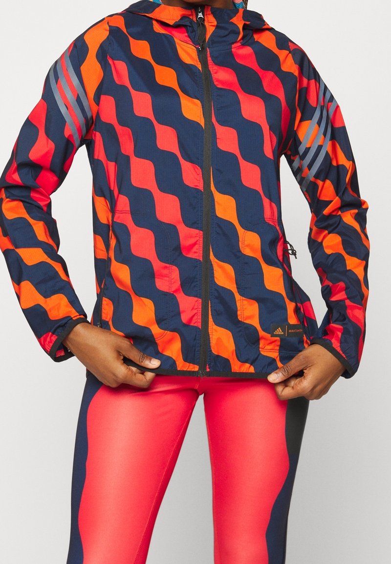 Personne portant une veste bleu marine avec des rayures ondulées orange et rouges et un legging de sport assorti rouge et bleu marine, ajustant l'ourlet de la veste.