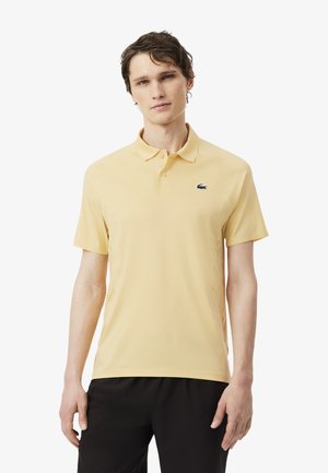 Giovane uomo con capelli castani corti, che indossa una polo gialla chiara e pantaloni neri, in piedi contro uno sfondo bianco.