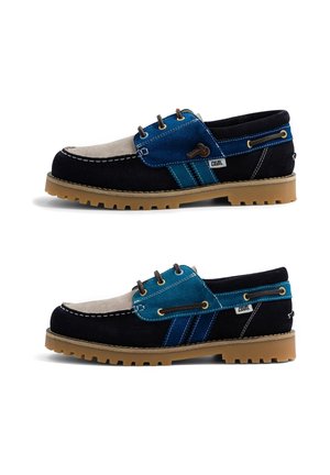 Personne portant des chaussures bateau bleues, beiges et noires avec des chaussettes blanches, assise sur un bateau à côté d'une marina avec des bateaux amarrés et de l'eau en arrière-plan.