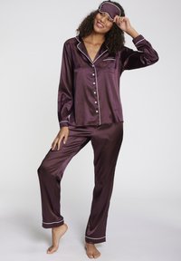 Purple satin pajama set with white piping, featuring a button-up shirt and long pants, accompanied by a matching sleep mask. Smooth texture.