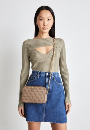 Frau trägt einen olivgrünen, gerippten Pullover mit Ausschnitt an der Brust, blauen Jeansrock und beige gemusterte Umhängetasche mit goldener Kettenriemen.