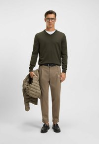 Maglione verde a scollatura a V, pantaloni beige, cintura nera e mocassini neri. Giacca leggera trapuntata tenuta a mano, caratterizzata da una texture liscia.