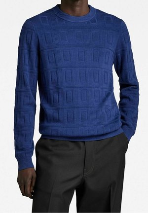 Homme portant un pull en maille texturée bleu avec des motifs géométriques et un pantalon noir, main dans la poche, sur un fond uni.