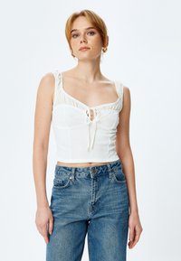 Biały top w stylu crop z teksturowanej tkaniny, kwadratowym dekoltem i falbaniastymi ramiączkami. Posiada detal w postaci zawiązania z przodu i jest zestawiony z niebieskimi dżinsami.